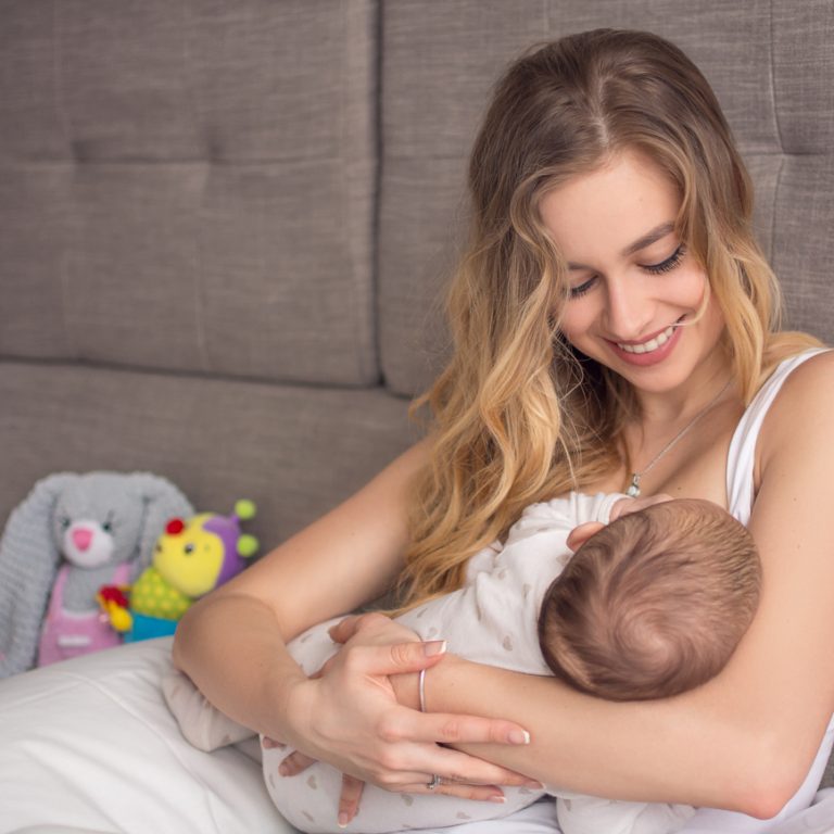 Breastfeeding baby. Pretty mother holding her newborn child. Mom smile and nursing infant. Beautiful woman and new born love at home. Blond mother breast feeding baby.